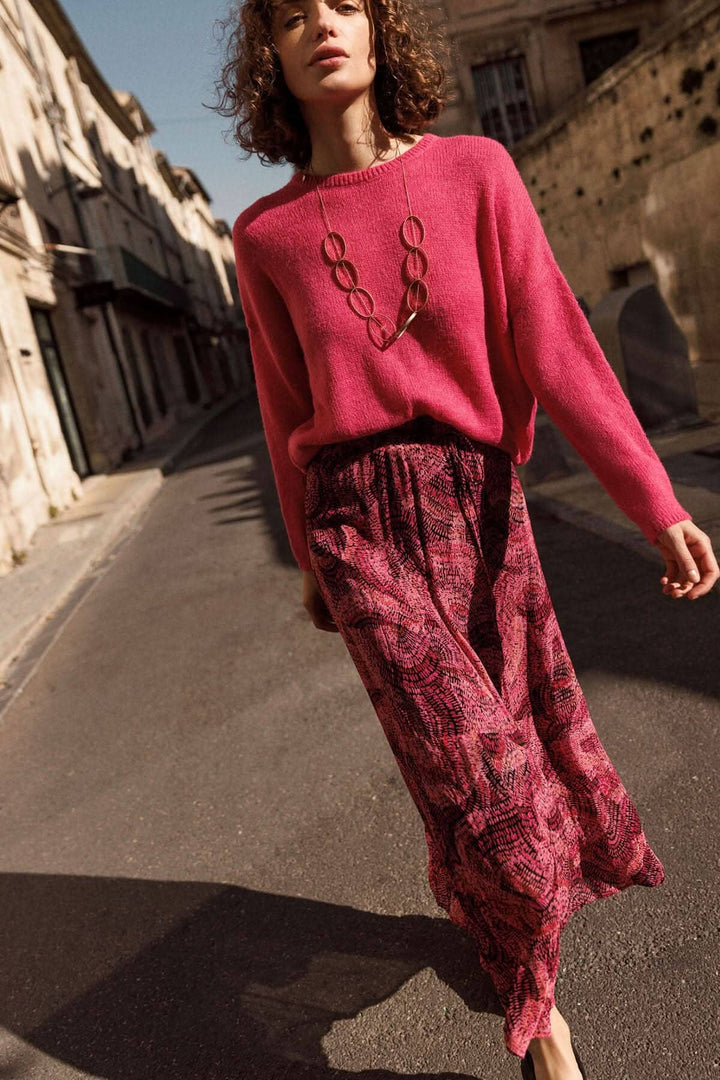 Femme portant une jupe longue abstraite et un pull rose, déambulant dans une rue ensoleillée.