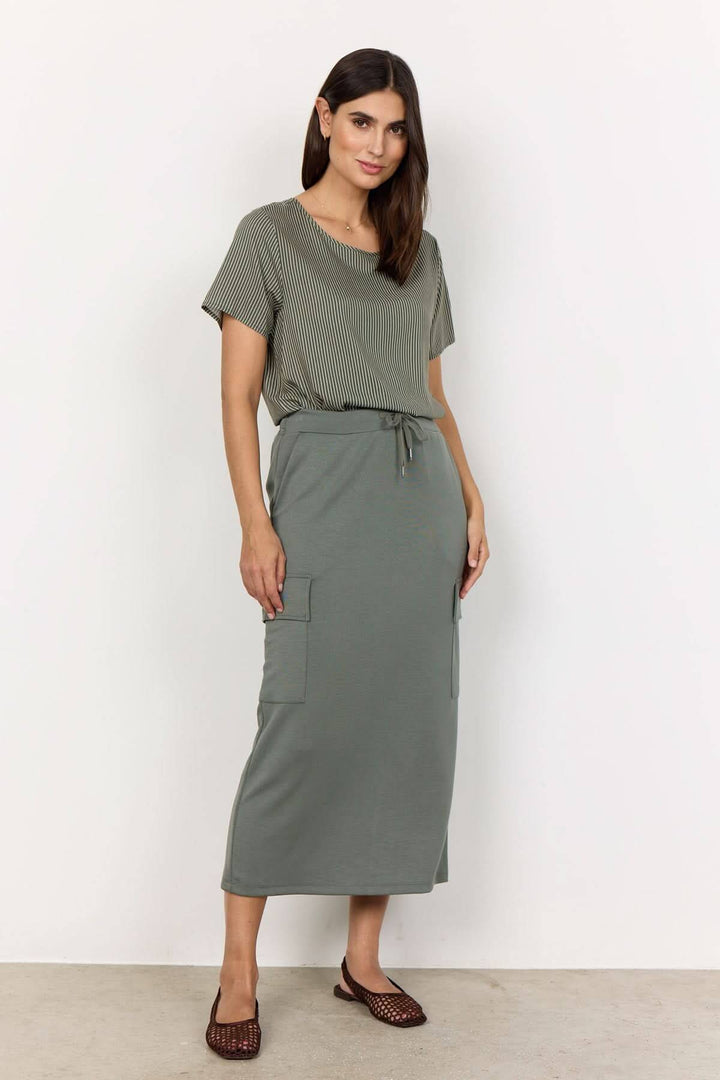 Femme portant une jupe longue souple verte avec poches, t-shirt assorti et sandales, style décontracté chic.