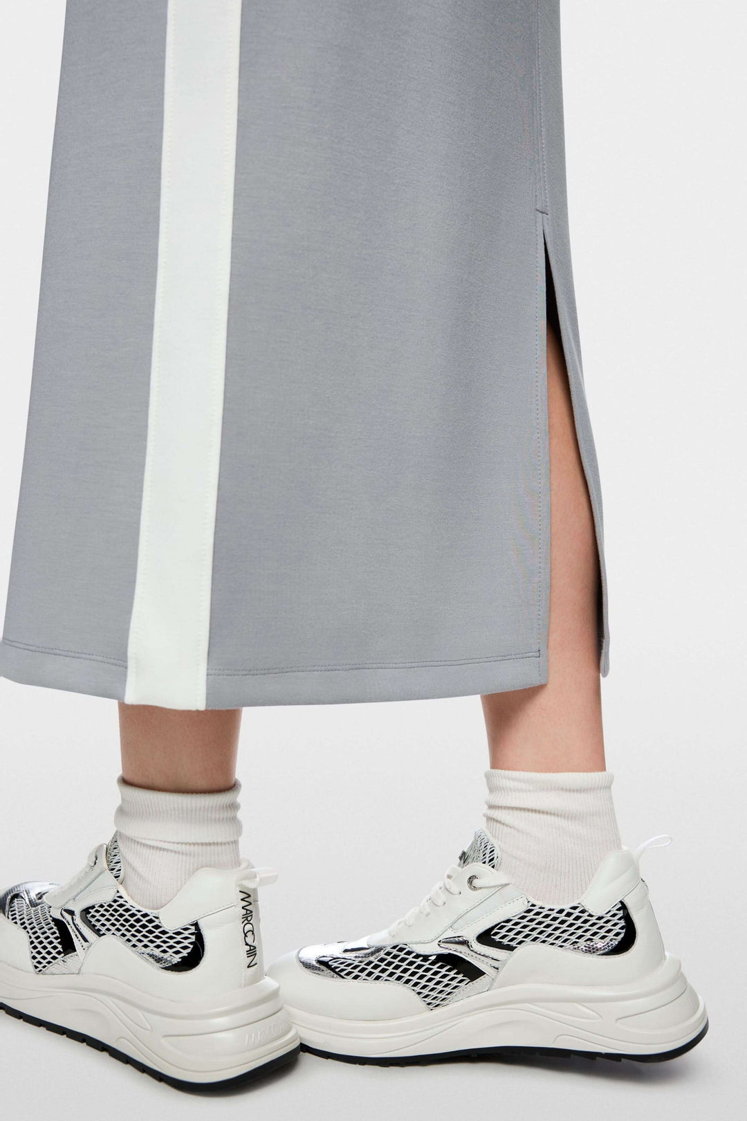 Détail de la jupe midi sportive avec fente à l'arrière, bandes contrastées et baskets blanches, logo Marc Cain visible.
