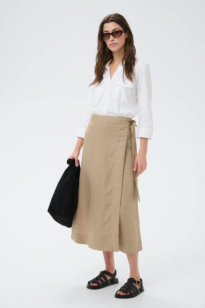 Femme portant la jupe porte-feuille Arbella beige, s'harmonisant avec une chemise blanche, offrant une allure décontractée et élégante.