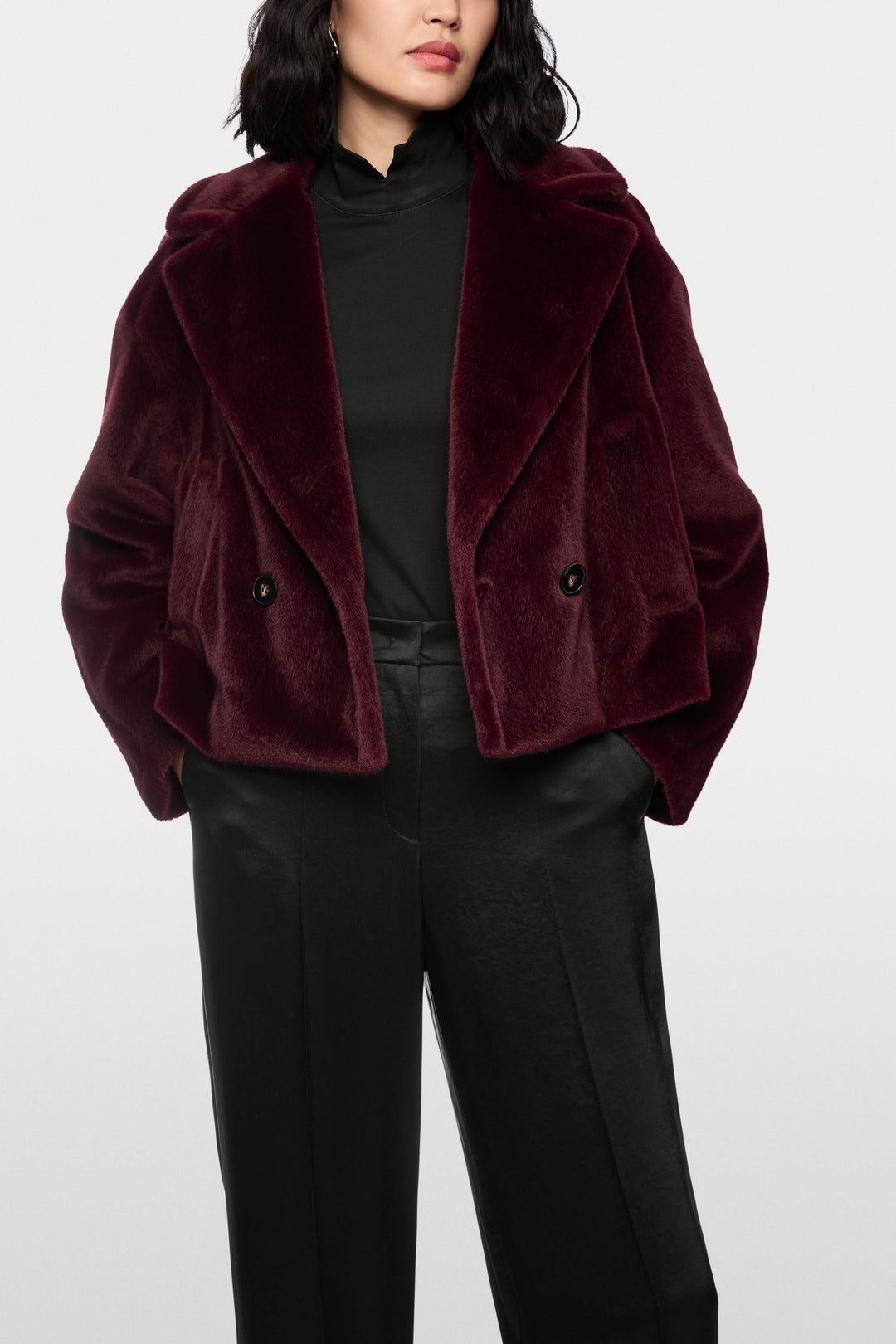 Manteau court en fausse fourrure bordeaux avec col tailleur, porté sur un haut noir et un pantalon noir.