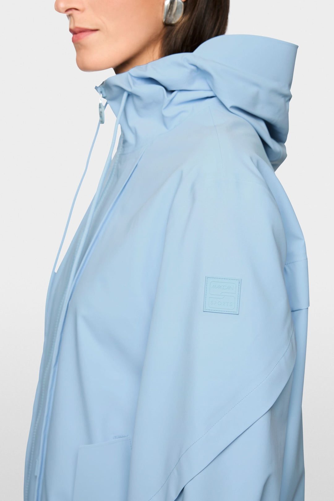 Femme portant un manteau d'extérieur léger avec capuche, couleur bleu ciel, avec fermeture à glissière et coupe épurée.
