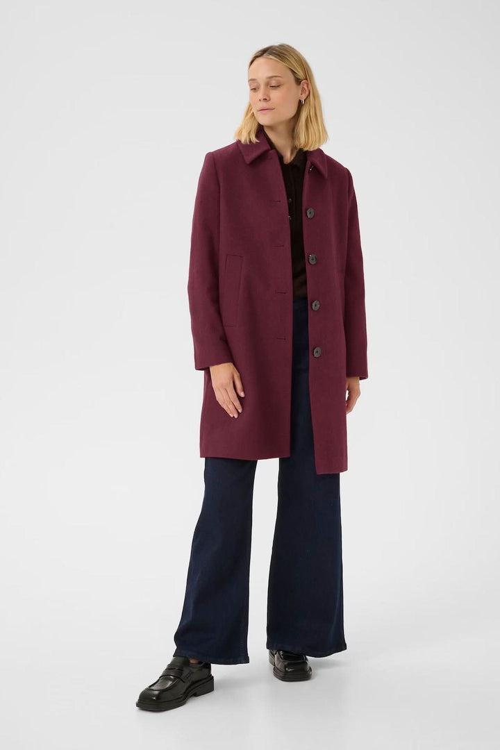 Femme portant un manteau en laine bordeaux à coupe droite avec col chemise et fermeture boutonnée, longueur mi-cuisse, sur un pantalon foncé.