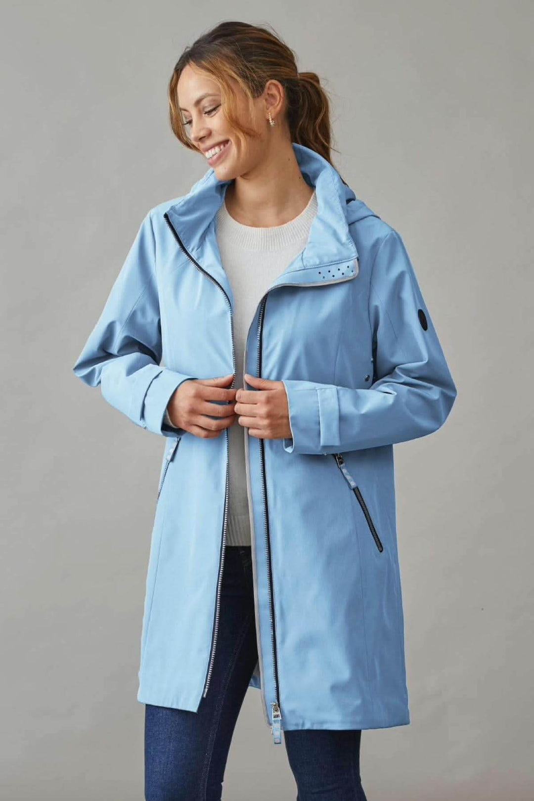Manteau long à capuchon bleu avec deux poches zippées et col ajustable, porté par une femme souriante.