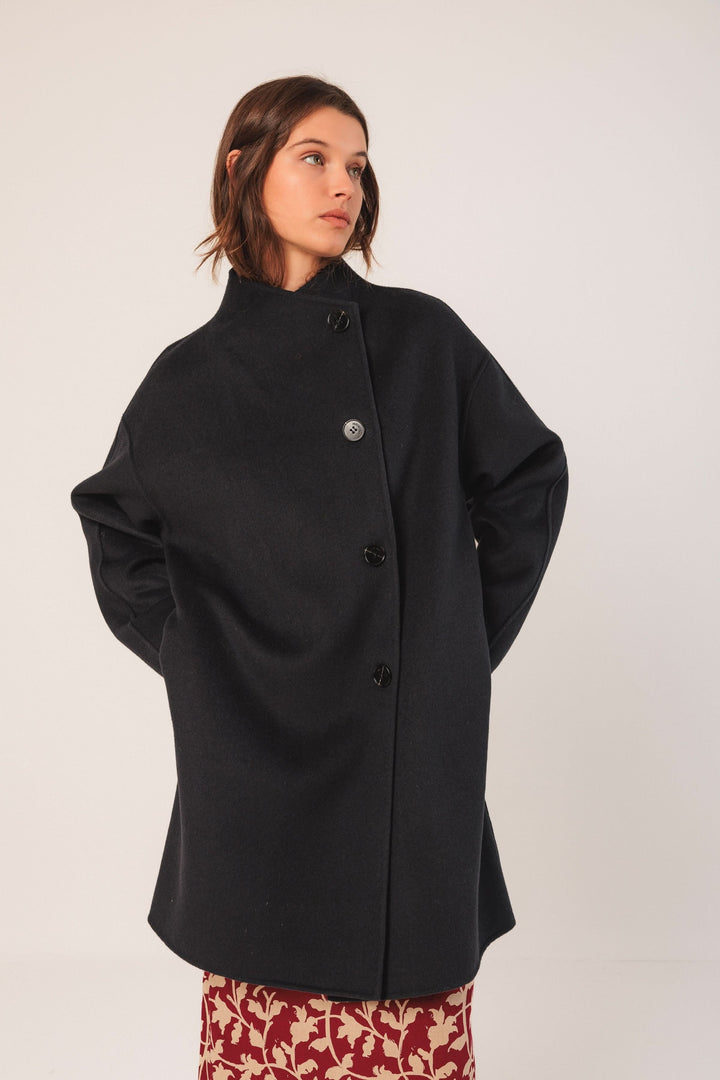 Manteau minimaliste noir avec coupe ample, col montant et fermeture boutonnée, porté sur une jupe imprimée.
