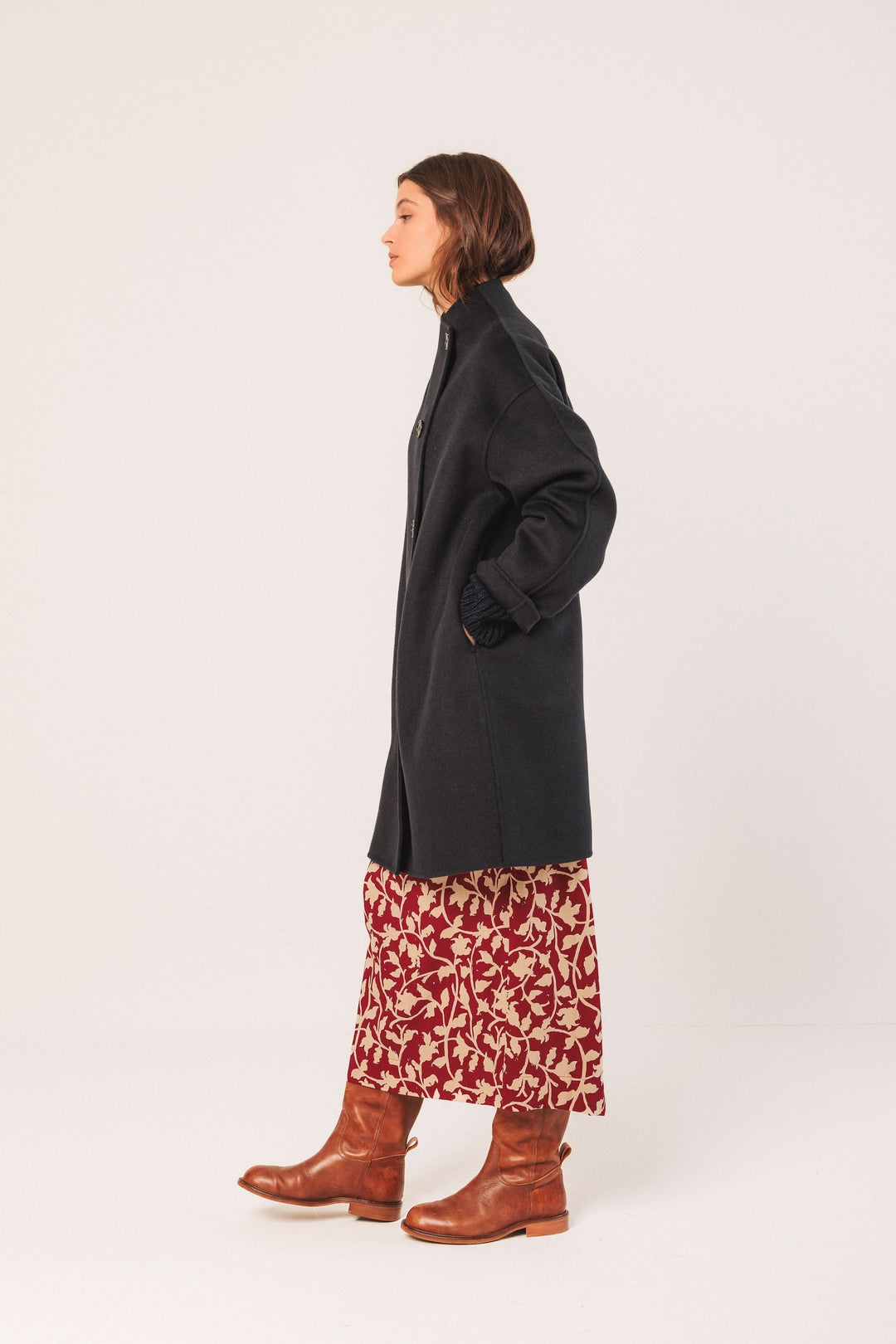 Manteau minimaliste noir avec col montant, porté sur une jupe florale et des bottes en cuir marron.