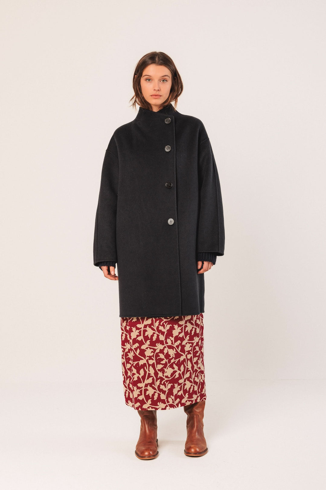 Manteau minimaliste noir à coupe ample et col montant, porté sur une robe florale avec des bottes en cuir.