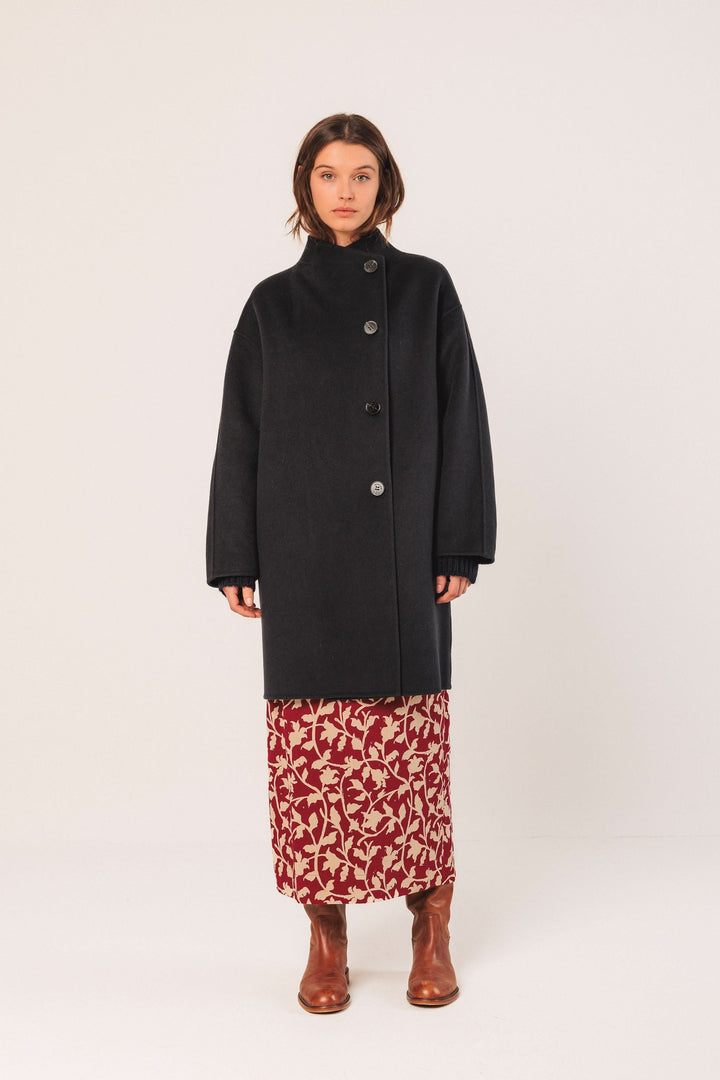 Manteau minimaliste noir à coupe ample et col montant, porté sur une robe florale avec des bottes en cuir.