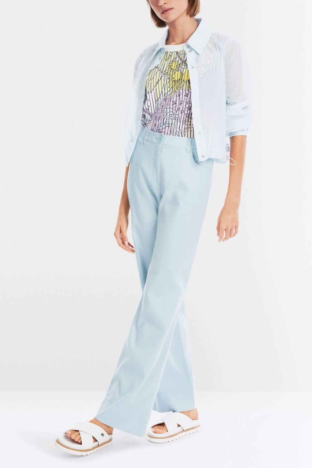 Femme portant un pantalon FAID bleu clair, top multicolore, avec une chemise légère et des sandales blanches.