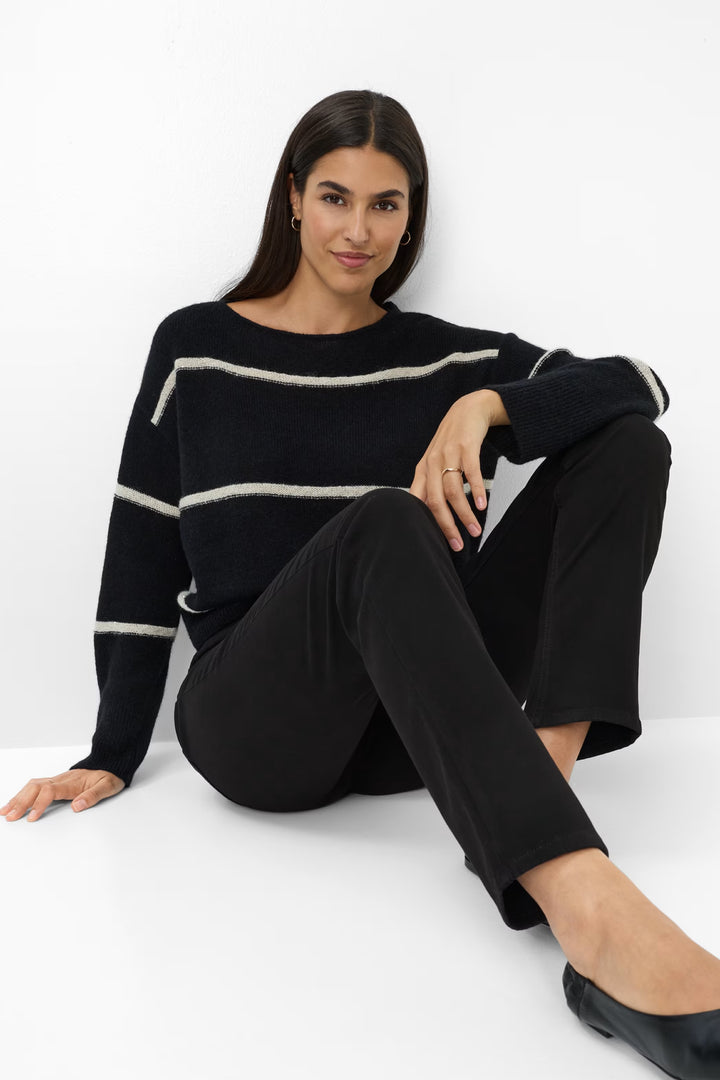 Femme portant un chandail rayé noir et beige avec un pantalon classique, assise sur un fond blanc.