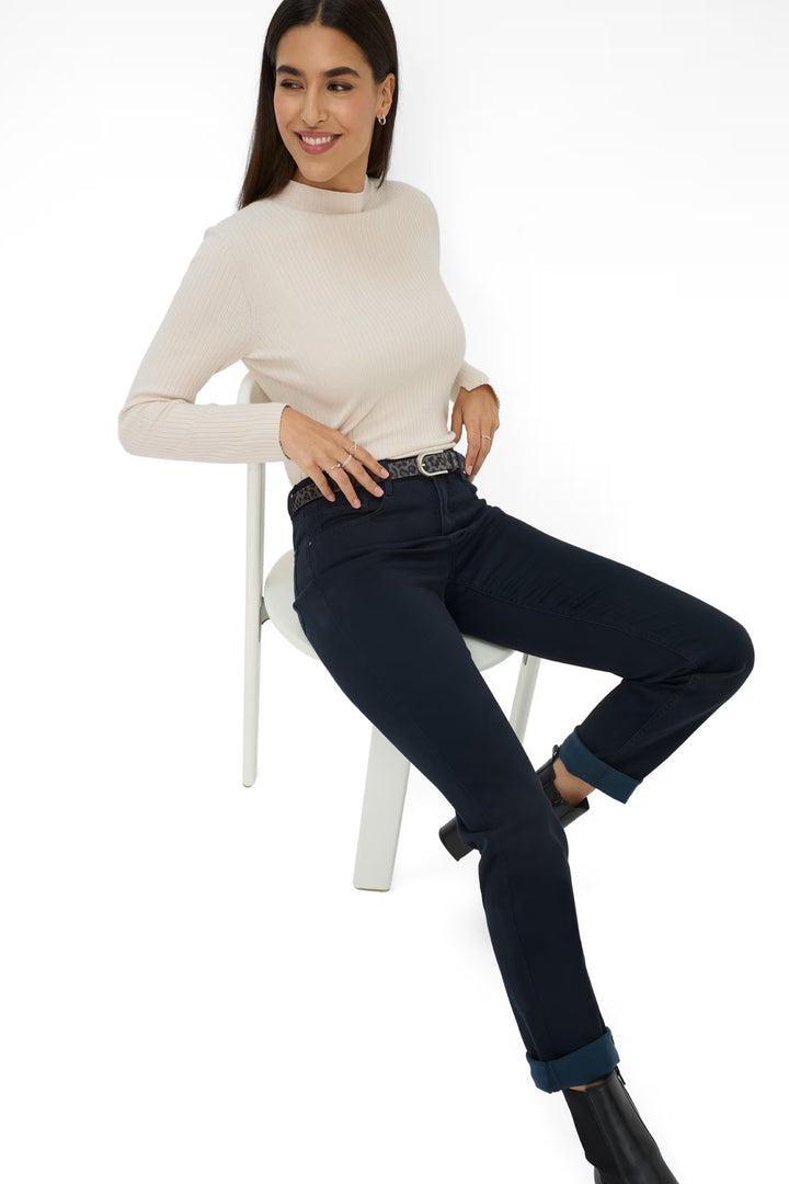 Femme portant le pantalon Mary en denim avec un chandail crème, assise sur une chaise blanche.