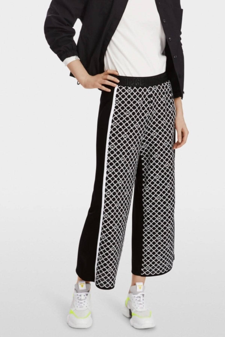 Pantalon WUSU noir et blanc à motif de chaînes, avec ceinture élastique, porté avec des baskets blanches.