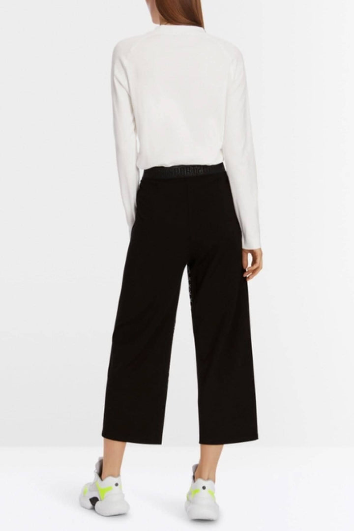 Pantalon WUSU large en mélange de matières, avec ceinture élastique, porté avec un pull blanc et des chaussures colorées.