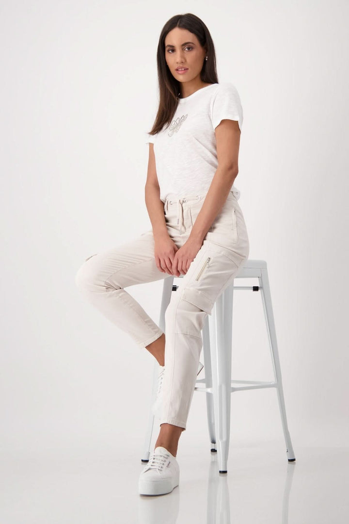 Femme portant un pantalon cargo beige avec t-shirt blanc, assise sur une chaise. Style décontracté et moderne.