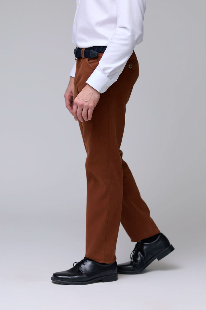 Homme portant un pantalon chino moderne marron avec coupe droite et tissu extensible, associé à une chemise blanche et des chaussures noires.