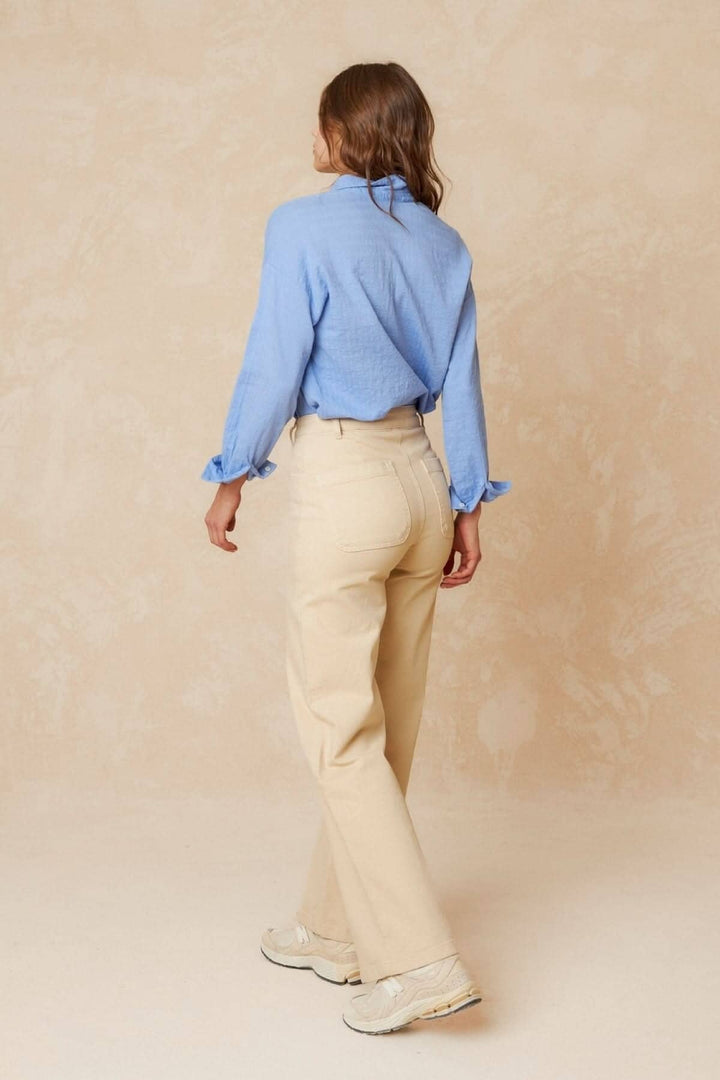 Femme portant un pantalon droit beige et une chemise bleue, vue de dos sur un fond beige.