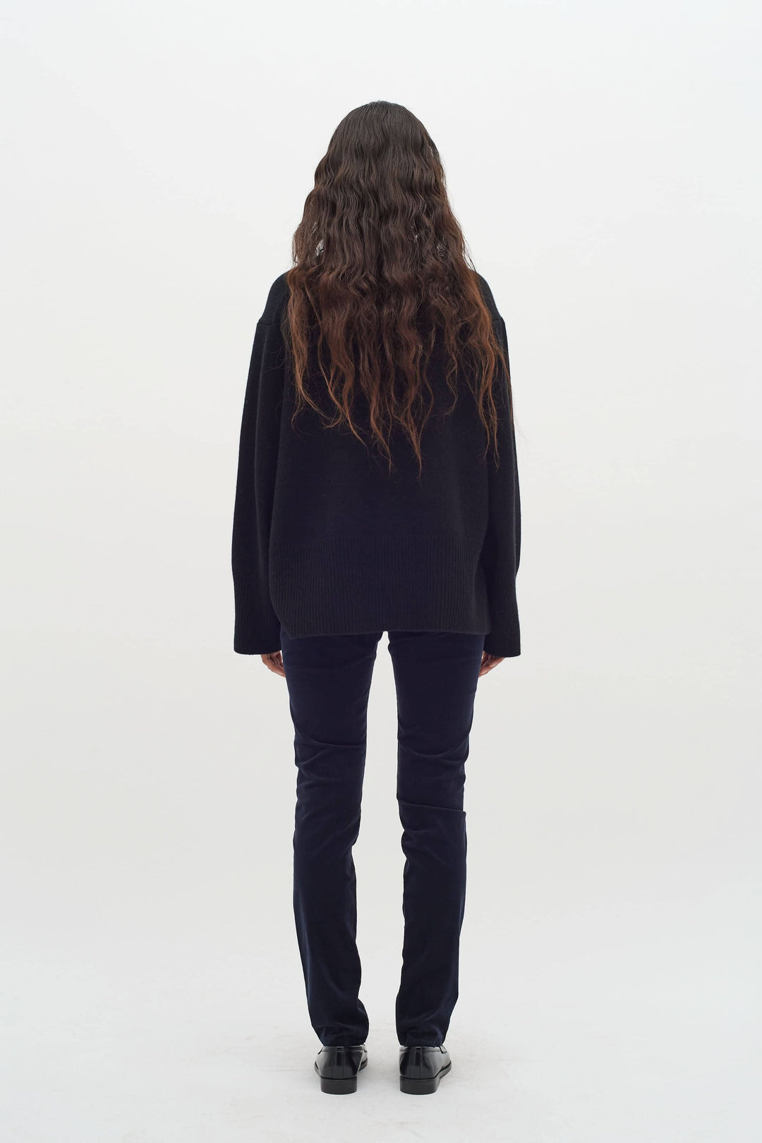 Femme de dos portant un pantalon en velours bleu marine et un pull noir, avec des cheveux longs et ondulés.