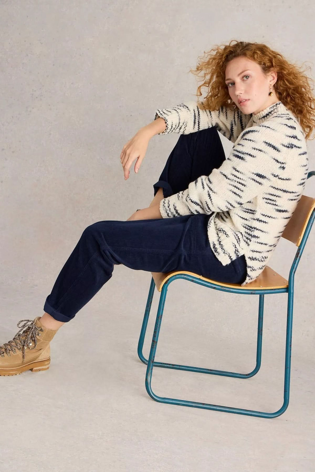 Femme assise sur une chaise avec un pantalon en velours côtelé noir et un pull à motifs, look décontracté.
