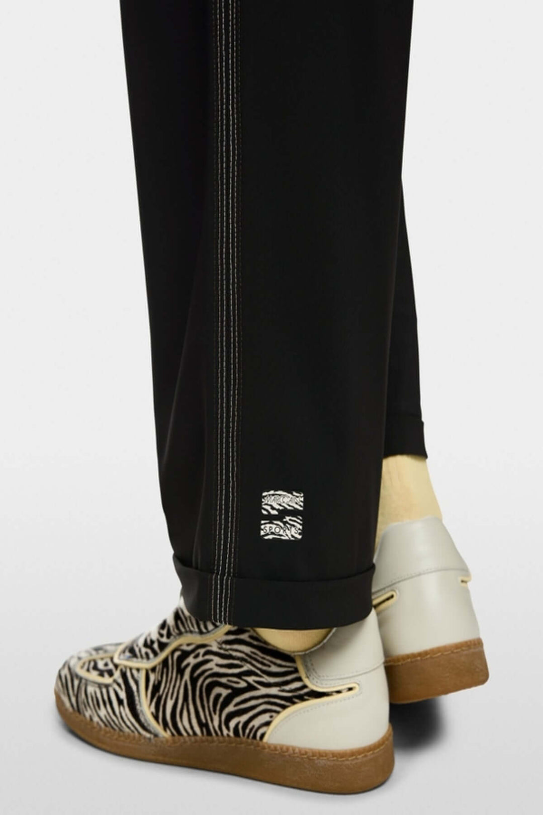 Gros plan sur le pantalon léger WUXI noir avec ourlet retourné et bande latérale, porté avec des chaussures à motif zébré.