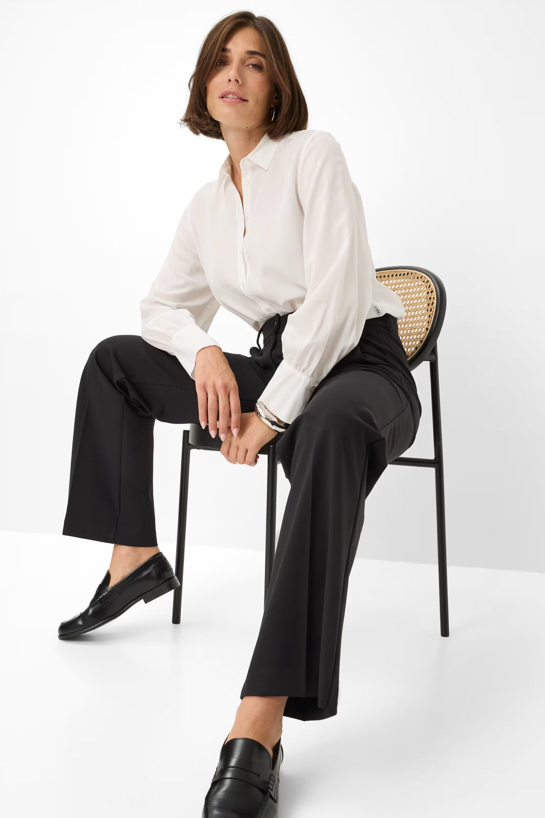 Femme assise sur une chaise, portant un pantalon palazzo noir et une chemise blanche, style chic et élégant.