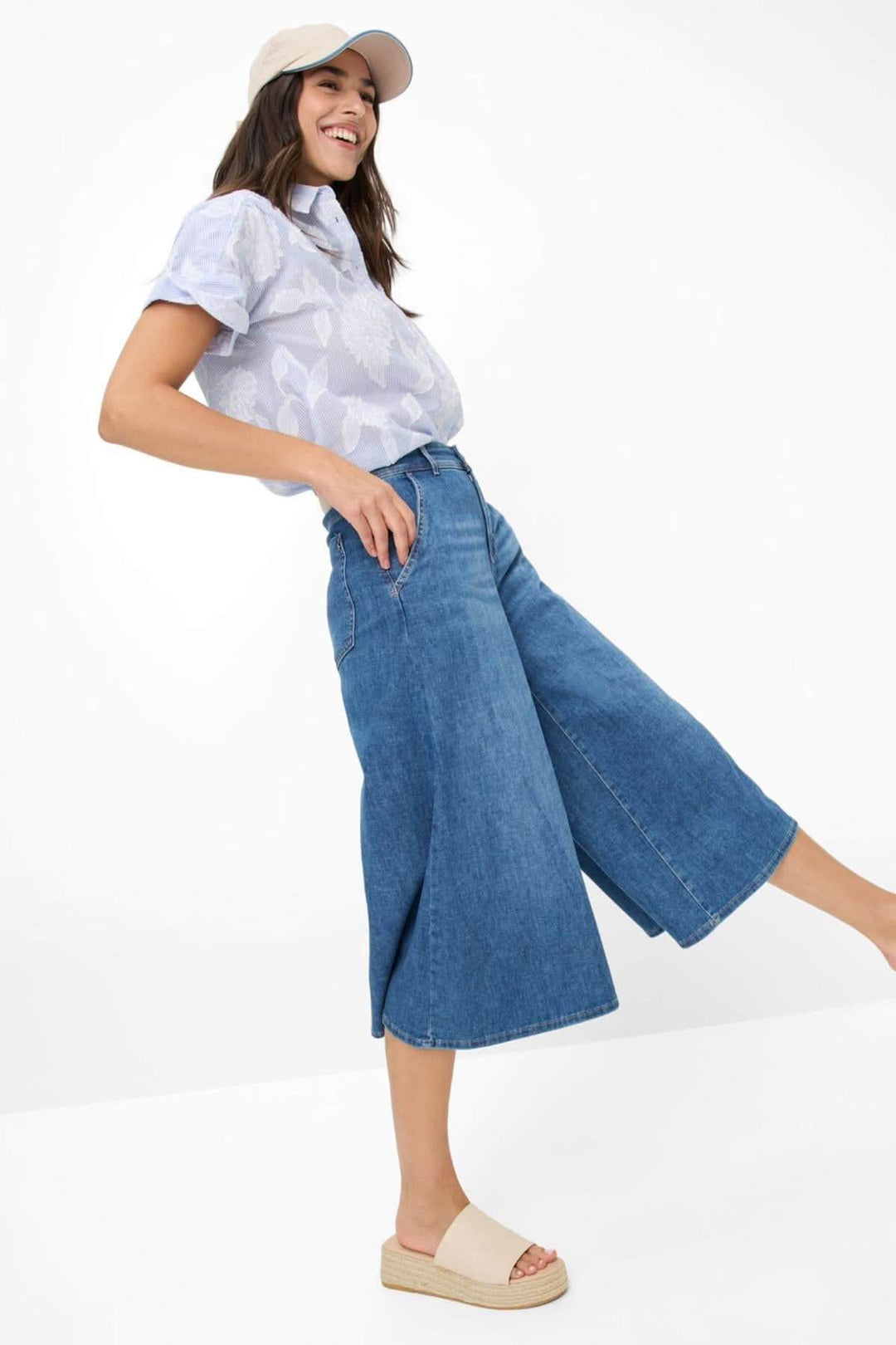 Femme souriante portant un pantalon palazzo bleu avec un haut à motifs et une casquette, dans une pose dynamique.