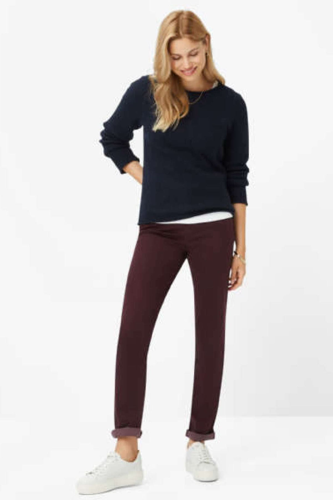 Femme souriante portant le pantalon velouté MARY en bordeaux avec un pull noir, stylisé avec des baskets blanches.