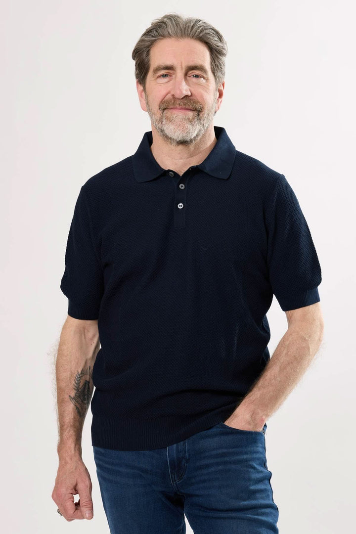 Homme souriant portant un polo raffiné bleu marine avec col structuré et manches courtes, style décontracté chic.