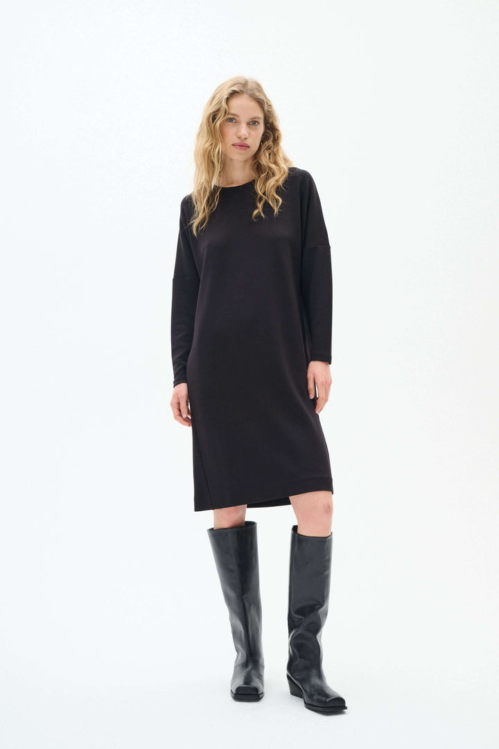 Robe Annie à manches longues, noire, avec silhouette fluide et encolure ronde, portée avec des bottes noires.