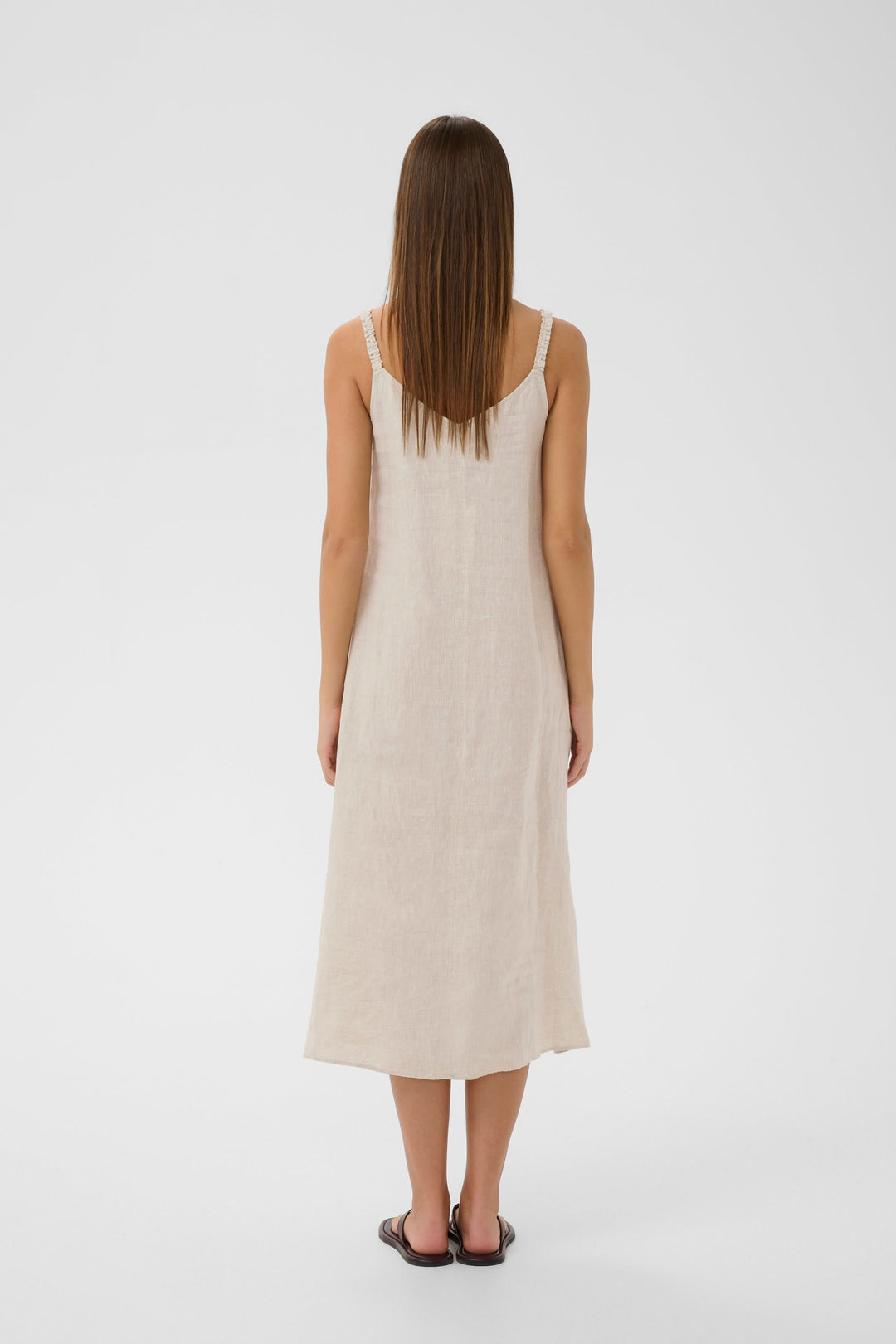 Femme portant la Robe Elinor midi fluide à bretelles fines en lin beige, vue de dos, montrant sa coupe ample et ses fines bretelles.