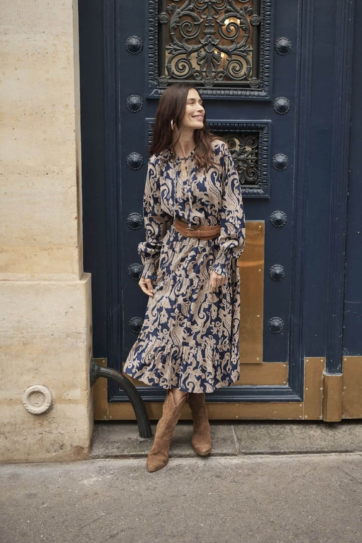 Femme souriante portant une robe longue beige et bleue, manches longues, devant une porte bleue.