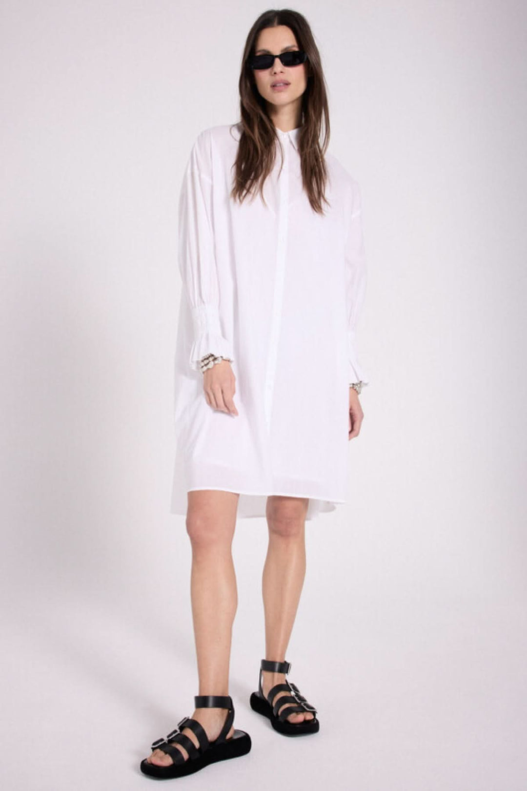 Robe chemise blanche avec détails plissés et cœur brodé, portée avec des sandales noires.