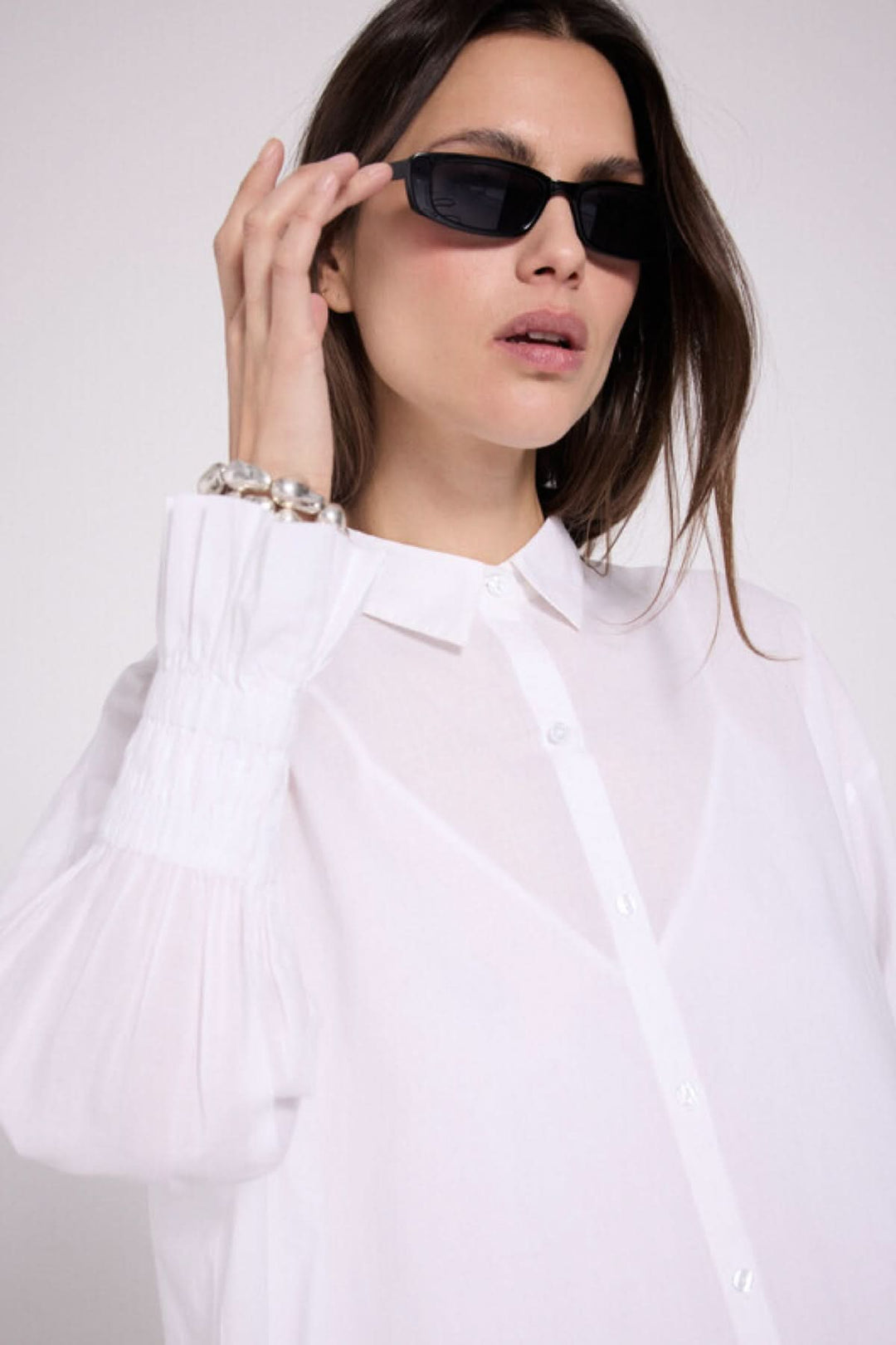 Femme portant une robe chemise blanche avec des manches bouffantes et des lunettes de soleil noires, affichant un look élégant.