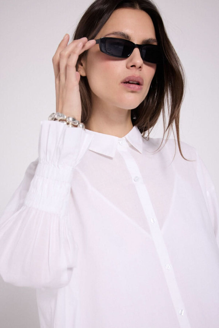 Femme portant une robe chemise blanche avec des manches bouffantes et des lunettes de soleil noires, affichant un look élégant.