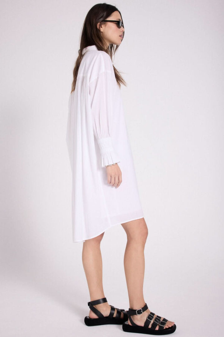 Robe chemise blanche avec détails plissés au dos, petit cœur brodé et manches longues, portée avec sandales noires.