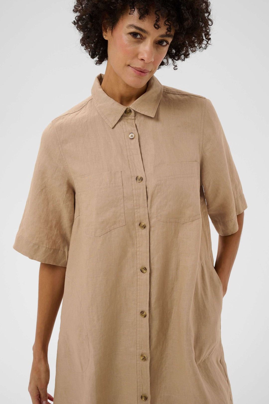 Femme portant une robe chemise en lin Sardiae beige avec manches courtes et fermeture boutonnée, présentant une coupe fluide et épurée.