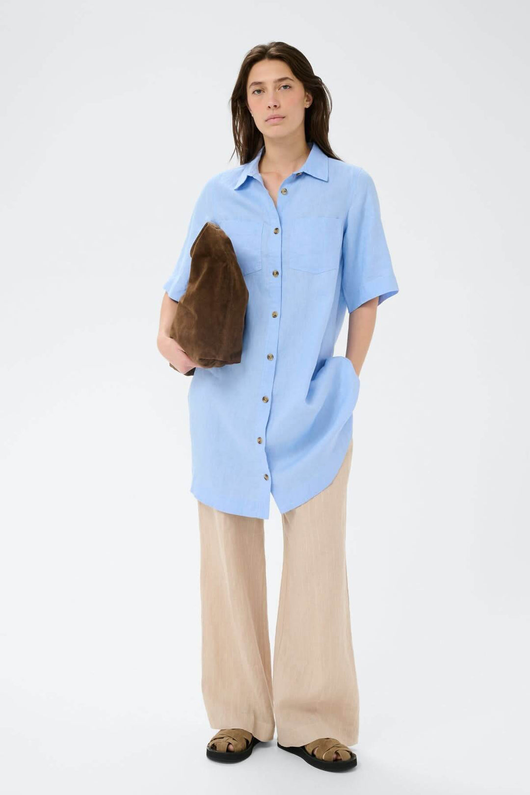 Femme portant une robe chemise en lin Sardiae, silhouette droite, couleur bleu. Boutonnée avec sac marron, look décontracté.