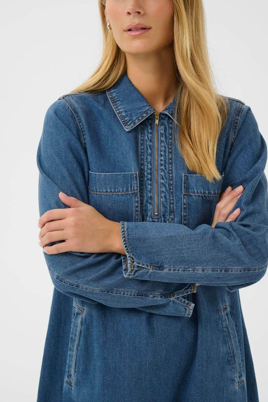 Femme portant une robe en denim, coupe droite, col chemise avec patte zippée, longueur au-dessus du genou, style décontracté et moderne.