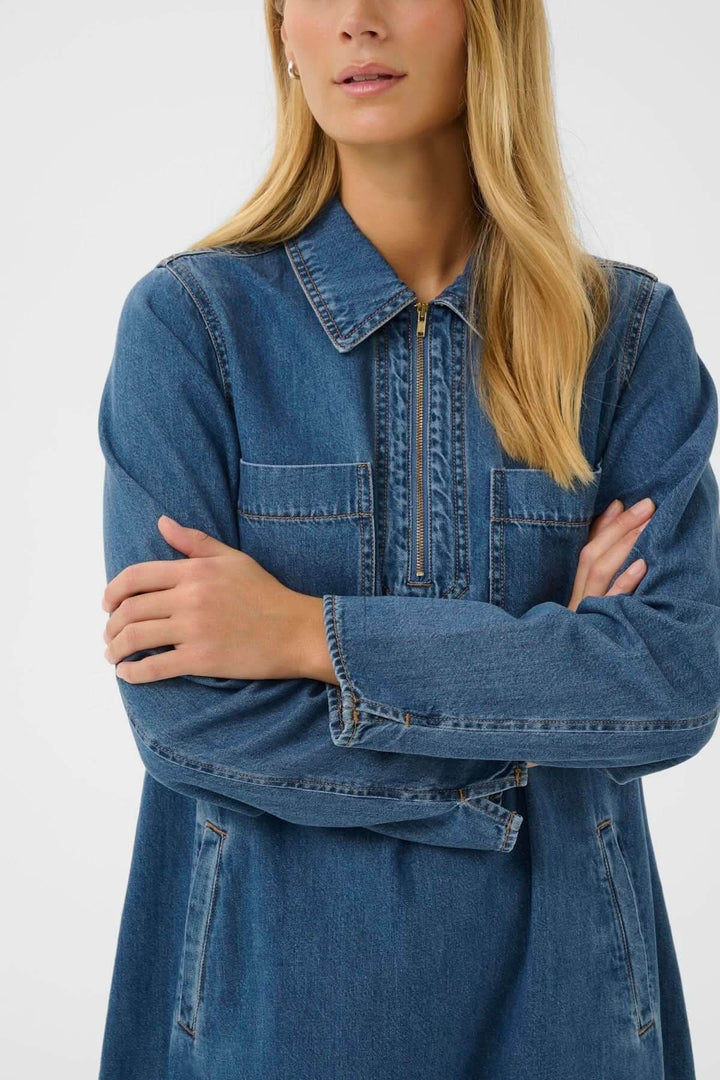 Femme portant une robe en denim, coupe droite, col chemise avec patte zippée, longueur au-dessus du genou, style décontracté et moderne.