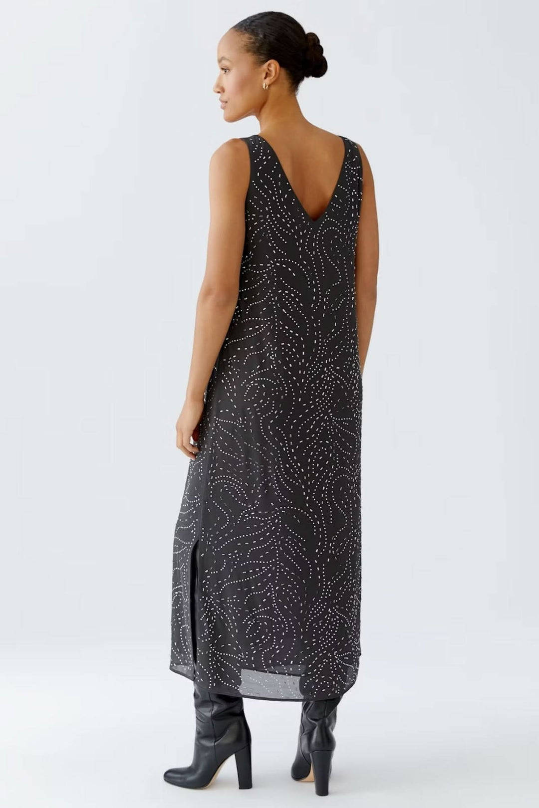 Femme portant une robe longue sans manches à imprimé pointillés, encolure en V, coupe fluide et fentes latérales, avec des bottes noires.