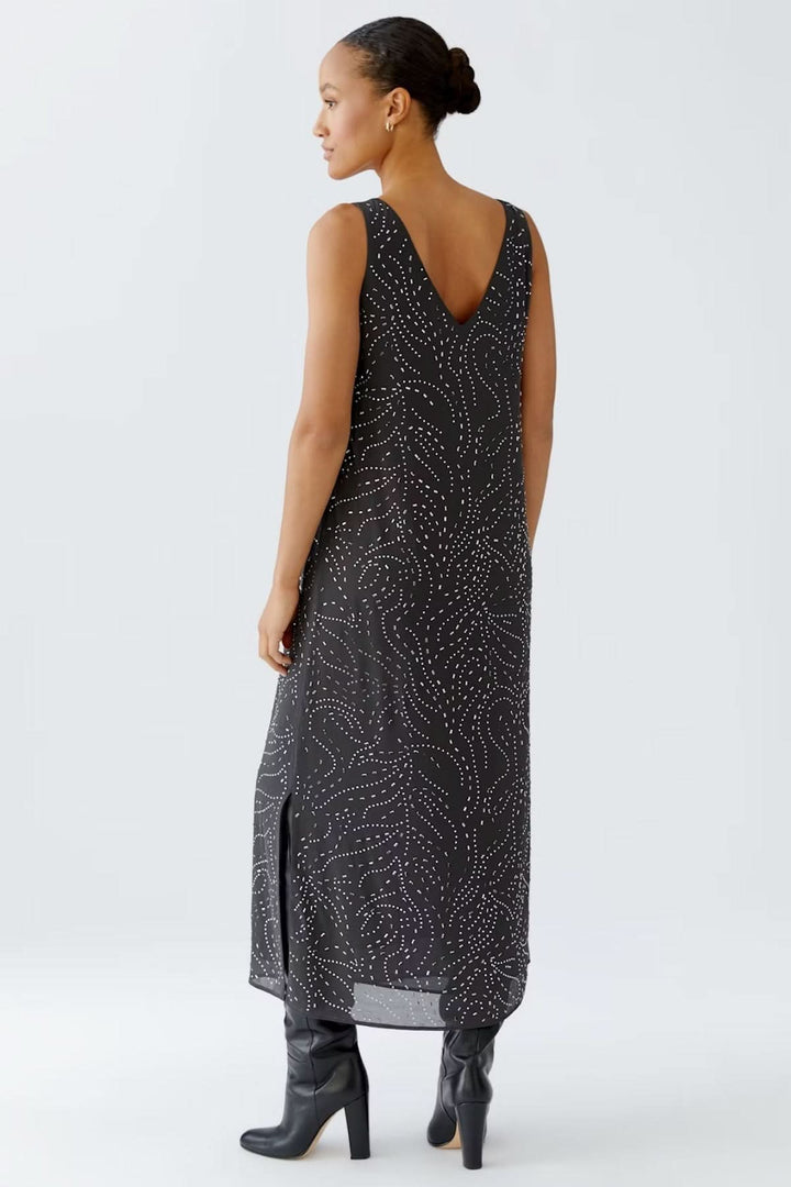 Femme portant une robe longue sans manches à imprimé pointillés, encolure en V, coupe fluide et fentes latérales, avec des bottes noires.