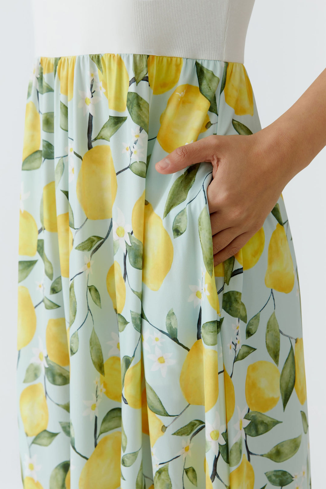 Gros plan sur une robe longue sans manches à jupe fluide avec motif citron, main dans la poche, tissu léger et coloré.
