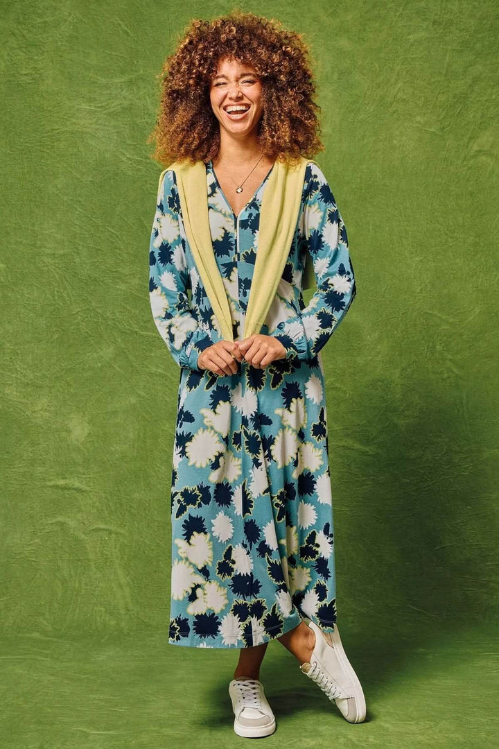 Femme souriante portant une robe mi-longue Megan à motifs floraux avec des baskets blanches et un gilet jaune.