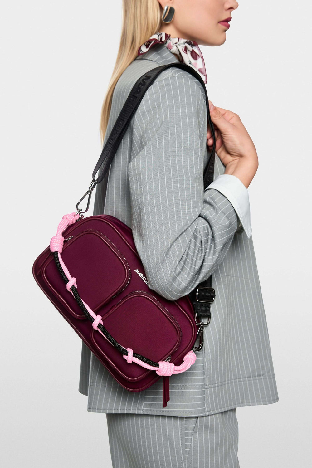 Sac bandoulière en néoprène avec poignée tressée et bandoulière ajustable, porté par une femme en costume gris.