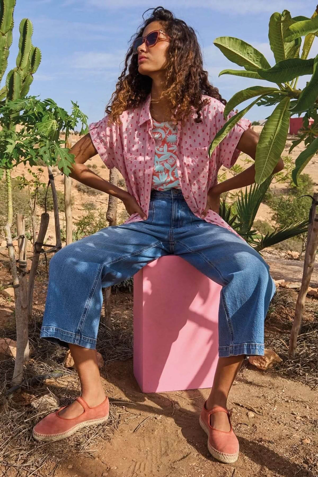 Femme assise sur un cube rose en tenue décontractée, t-shirt imprimé, jeans larges, cadre en plein air.