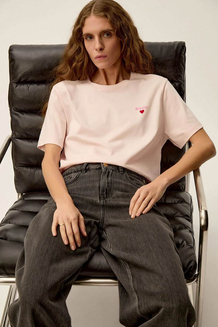Femme portant un t-shirt rose doux Gazoline avec imprimé cœur, assise sur une chaise moderne.