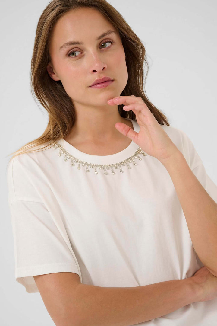 Femme portant un t-shirt blanc "Helen" à manches courtes et col rond, accessoirisé d'un collier élégant, montrant un look minimaliste.