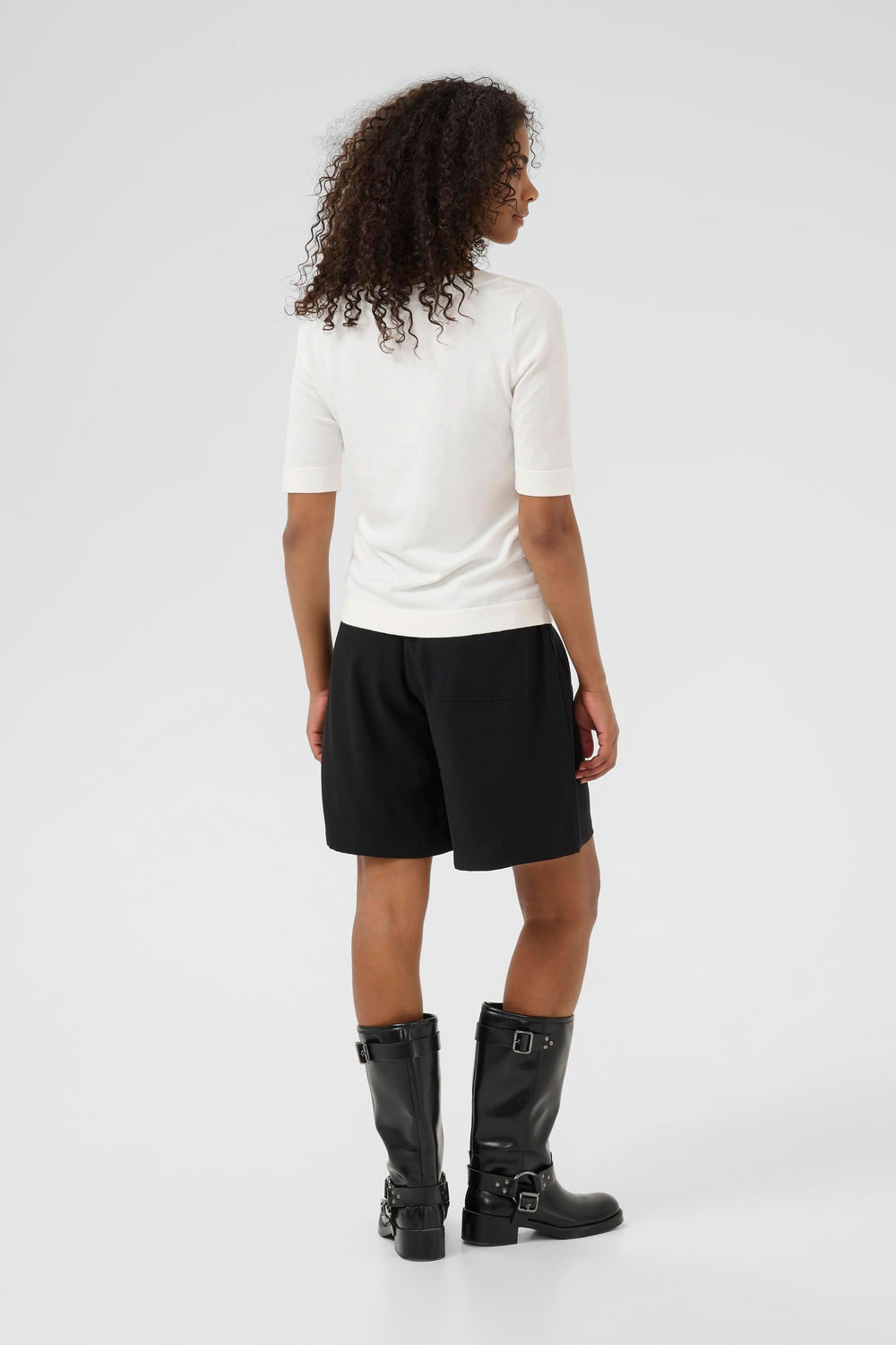 Femme portant un t-shirt blanc à manches mi-longues avec shorts noirs et bottes en cuir, vu de dos.