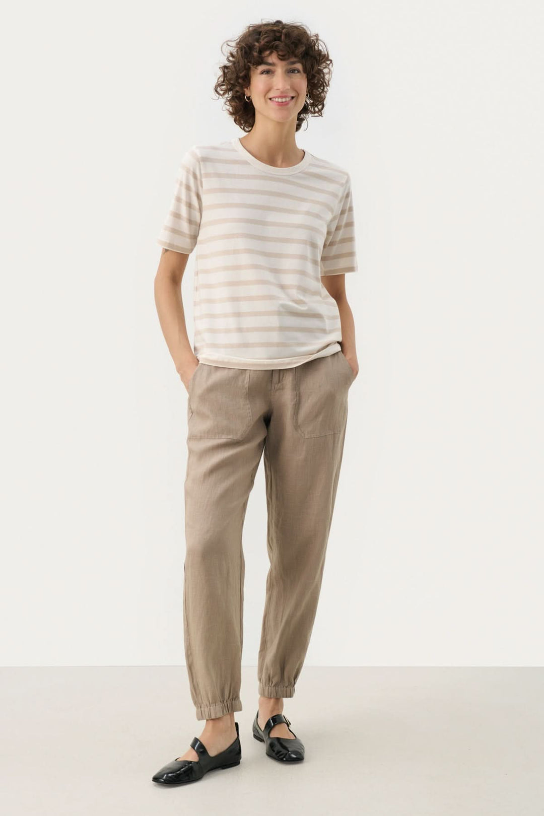 Femme souriante portant un t-shirt rayé et un pantalon beige sur un fond neutre.