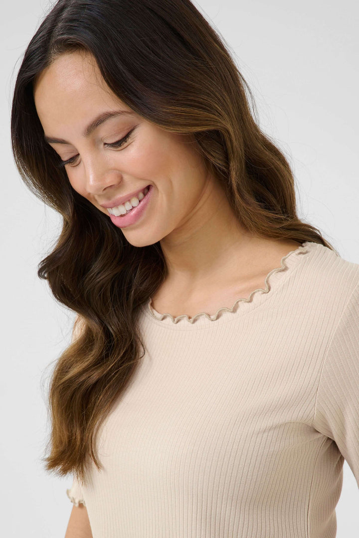 Femme souriante portant un T-shirt Ribba en jersey côtelé beige avec encolure à finition festonnée.