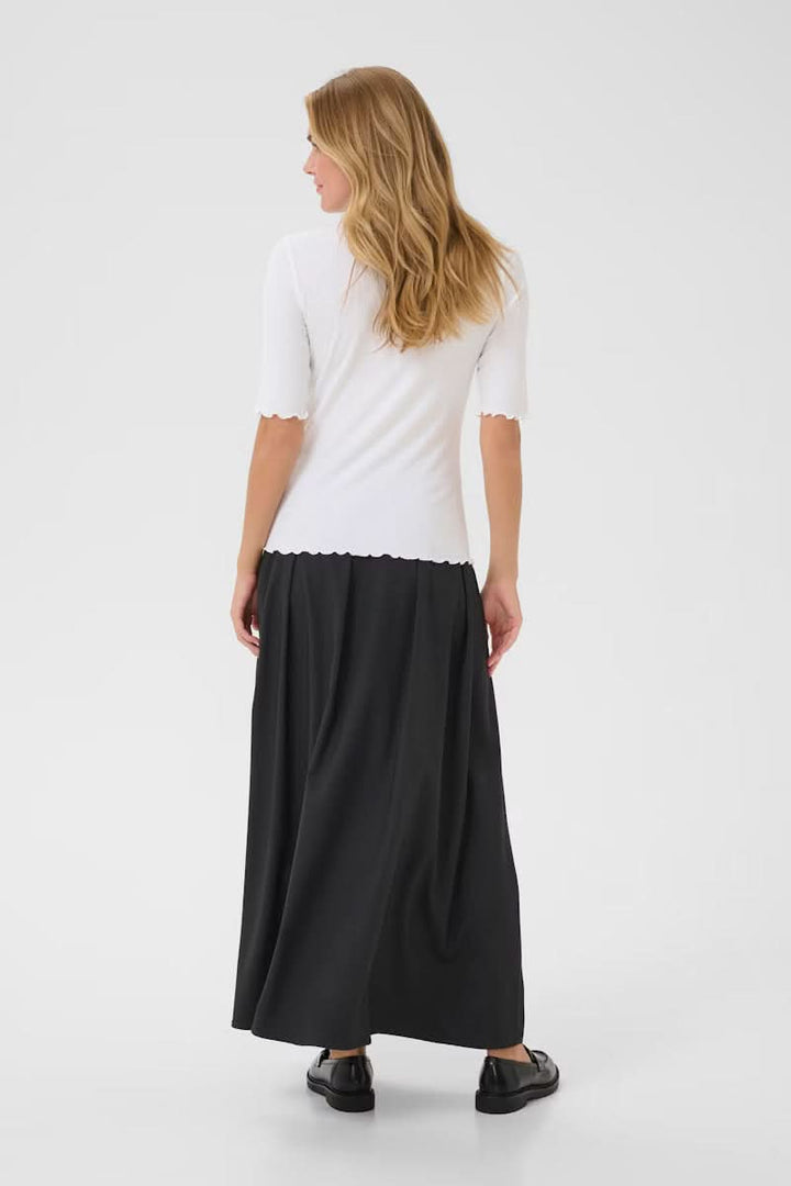 Femme portant un T-shirt Ribba blanc à col montant festonné et manches mi-longues, associé à une jupe longue, vue de dos.